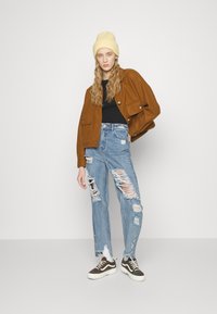 Brun ullblanding jakke med to frontlommer, svart topp, slitte lyseblå jeans og svart/sølv sneakers med hvite såler.