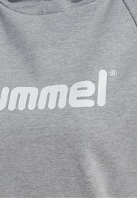 Tissu gris avec logo blanc « hummel » partiellement visible imprimé, montrant la texture du tricot du matériau.