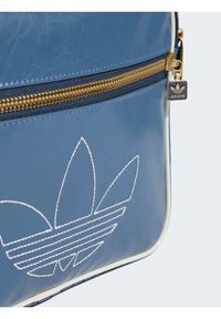 Blå syntetisk läderväska med en vit konturerad trefoil-logotyp. Den har en gyllene dragkedja och en kontrasterande vit kant runt kanterna.