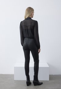 Zwarte doorschijnende blouse met geplooide mouwen, gecombineerd met aansluitende zwarte leggings met een textuur van bloempatronen en zwarte enkellaarsjes.