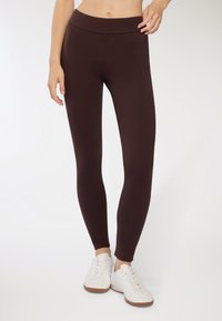 Brązowe legginsy wykonane z elastycznego, gładkiego materiału z szerokim pasem w talii. Noszone z białymi trampkami, o niskim stanie i długości do kostek.