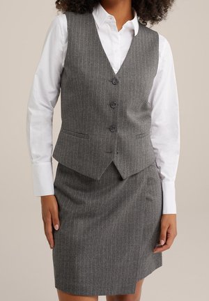 Kvinde iført en grå striped vest og matchende nederdel over en hvid skjorte med knapper, stående med hænder afslappet ned langs siden.