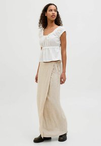 Bela bluzica s kratkimi rokavi in nabrano podrobnostjo, v kombinaciji z bež ovito krilo, zavezanem v pasu. Model nosi črne škornje.