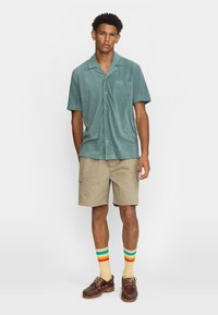 Turquoise shirt met korte mouwen en een zak, gecombineerd met beige cargoshorts. Draagt met veelkleurige gestreepte sokken en bruine schoenen.