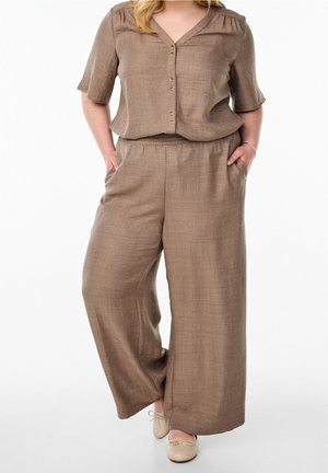 Vrouw draagt losse bruine kortere mouw met knopen top en bijpassende wijde broek met beige ballerina's, handen in de zakken.
