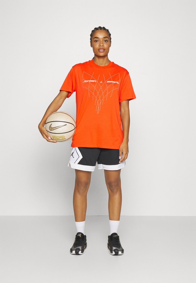 T-shirt de sport orange avec un design graphique, shorts noirs avec des bords blancs et baskets noires, tenant un ballon de basket beige avec un logo.