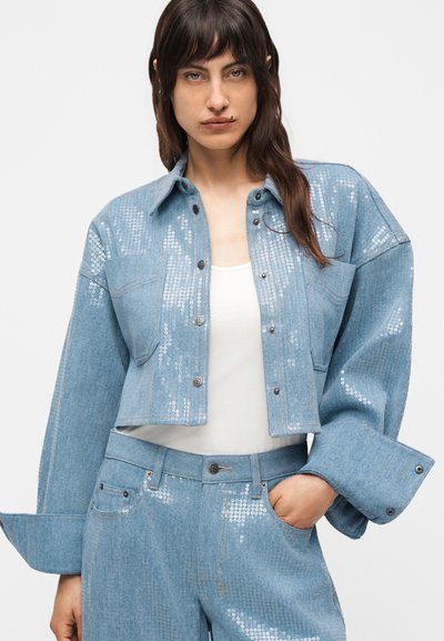 Νεαρή γυναίκα με ανοιχτό γαλάζιο cropped τζιν μπουφάν και αντίστοιχο παντελόνι με ασημί παγιέτες, λευκό τοπ, στέκεται με το ένα χέρι στην τσέπη.