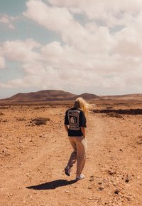 Osoba w czarnej graficznej koszulce i jasnych beżowych dresach spacerująca po szutrowej ścieżce w jałowym krajobrazie z odległymi górami pod pochmurnym niebem.