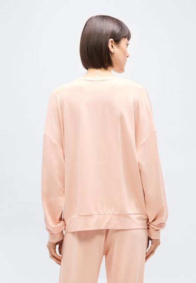 Γυναίκα με κοντά σκούρα μαλλιά, φοράει άνετο, ανοιχτό ροζ μακρυμάνικο τοπ και παντελόνι που ταιριάζει, εμφανίζεται από πίσω μπροστά σε ένα απλό φωτεινό φόντο.