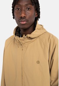 Beige letvægtsjakke med hætte, der har en lynlås foran og snore. Indeholder et subtilt logo nær brystet og en struktureret inderside.