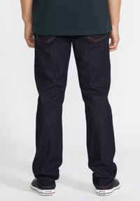 Jeans en denim bleu foncé avec une coupe droite, dotés de poches arrière et de coutures dorées contrastantes, associés à des baskets noires.