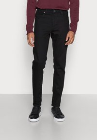 Jeans de mezclilla negra con corte slim, diseño de cinco bolsillos y cierre de botón. Combinados con zapatillas negras, en un fondo neutro.