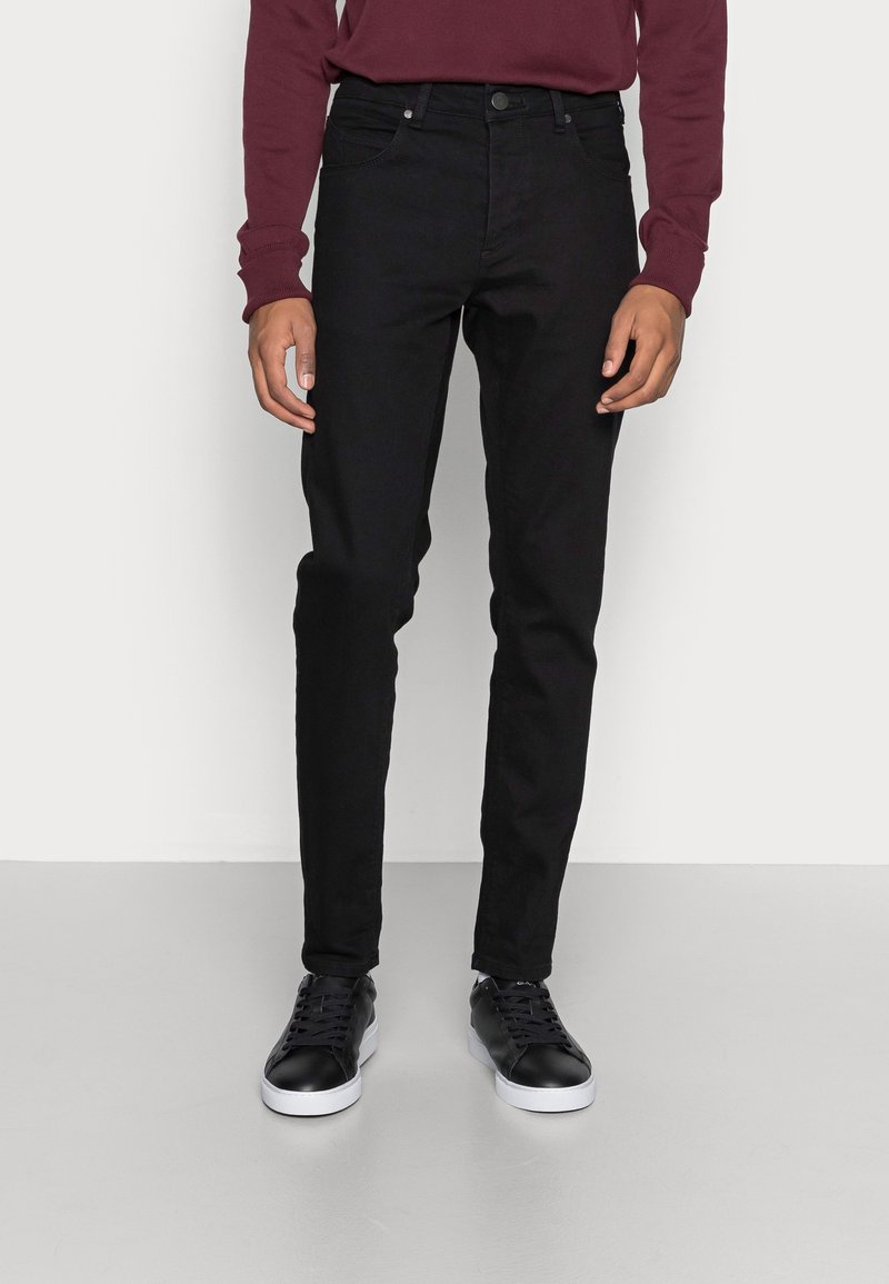 Jeans de mezclilla negra con corte slim, diseño de cinco bolsillos y cierre de botón. Combinados con zapatillas negras, en un fondo neutro.