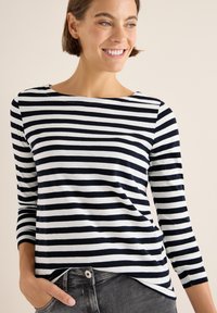 Langærmet stribet top i navy og hvid bomuld; har bådudskæring og skrå kant. Sammen med grå jeans, hænderne i lommerne.