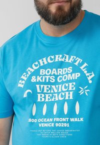 Blauwe katoenen T-shirt met witte grafische tekst, waaronder "BEACHCRAFT L.A." en "VENICE BEACH," met een relaxte pasvorm en korte mouwen.
