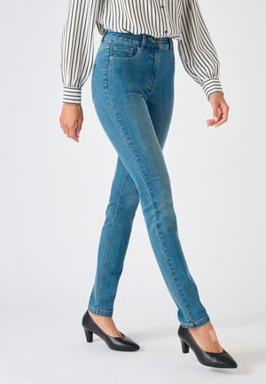 Personne portant un jean slim bleu, des talons noirs et un chemisier blanc à rayures verticales noires sur un fond blanc uni.