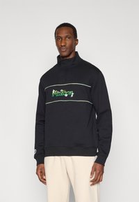 Μαύρο πουλοβεράκι με ψηλό γιακά, διακοσμημένο με πράσινο και λευκό κέντημα κειμένου, και αντίθετες ρίγες στο στήθος και τα μανίκια.