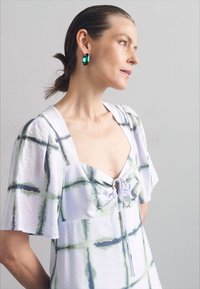Witte jurk met een blauw en groen ruitpatroon, korte fladdermouwen, nagekrompen halslijn met strikjes en een paar groene oorbellen.