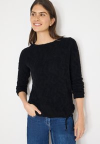 Maglione nero dalla texture morbida con maniche a 3/4, scollo tondo e dettagli con coulisse all'orlo. Abbinato a jeans blu.