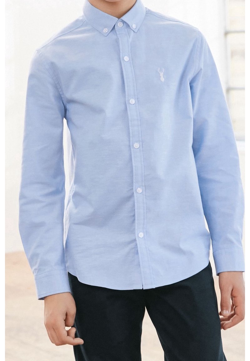 Garçon portant une chemise bleu clair à manches longues boutonnée avec un petit logo de cerf sur la poitrine et un pantalon noir.