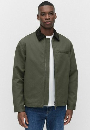 Veste vert olive avec un col en velours côtelé noir, fermeture éclair sur le devant et poche poitrine. Portée par-dessus une chemise blanche, associée à un jean bleu.