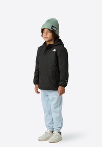 Veste noire imperméable avec capuche, poignets élastiques ; pantalon de survêtement bleu clair ; bonnet côtelé vert ; baskets blanches avec semelles texturées.