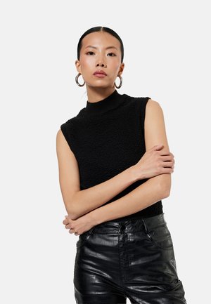 Vrouw met steil haar draagt een gestructureerde zwarte mouwloze top, grote oorringen en zwarte hoge taille nepleren broek, armen gekruist.