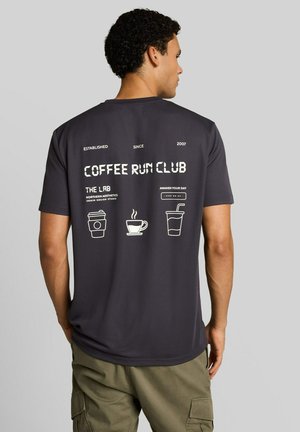 Man gezien van achteren, die een zwart T-shirt draagt met witte koffietekst en iconen met de tekst "Coffee Run Club" en drankillustraties.