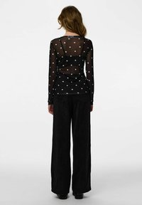 Czarna siateczkowa bluzka z długim rękawem w białe grochy, nałożona na czarną kamizelkę, w połączeniu z szerokimi czarnymi spodniami sztruksowymi.