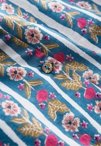 Tessuto blu con righe bianche, con motivi floreali in rosa e oro. Un piccolo bottone marrone è visibile tra i motivi.