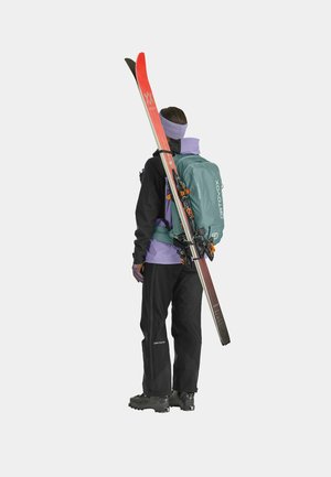 Ski equipment user, wearing a black jacket, purple shirt, and black pants, carries red and black skis and a teal backpack.
