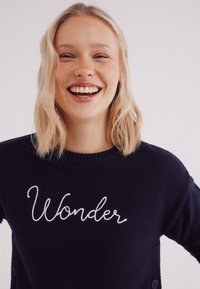 Marineblauwe trui met een ronde hals, voorzien van witte geborduurde tekst "Wonder" op de voorkant. Zachte textuur, met knoopaccenten op de mouwen.