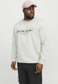 Crème sweatshirt met een zwart grafisch ontwerp, ronde halslijn en geribbelde manchetten. G gedragen met zwarte cargo-broeken en een roze pet.