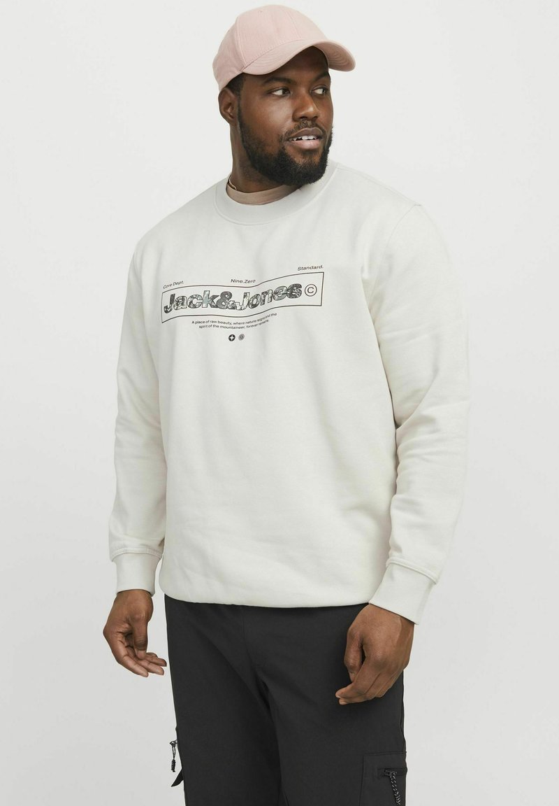 Crème sweatshirt met een zwart grafisch ontwerp, ronde halslijn en geribbelde manchetten. G gedragen met zwarte cargo-broeken en een roze pet.