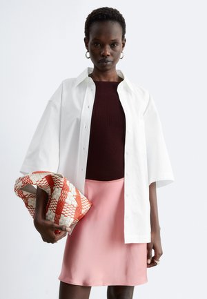 Femme portant une chemise blanche oversize à boutons, un haut côtelé foncé, une jupe rose, tenant une grande pochette tissée rouge et blanche.
