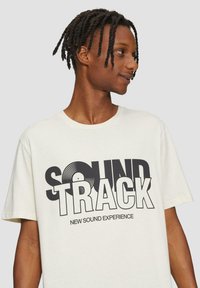 Crème-kleurige katoen t-shirt met een grote grafische print met de tekst "SOUND TRACK" en een afbeelding van een vinylplaat. Korte mouwen.