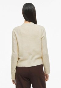 Žena s dugom tamnom kosom koja nosi kremasti džemper i tamno smeđe hlače, prikazana s leđa na jednostavnoj bijeloj pozadini.