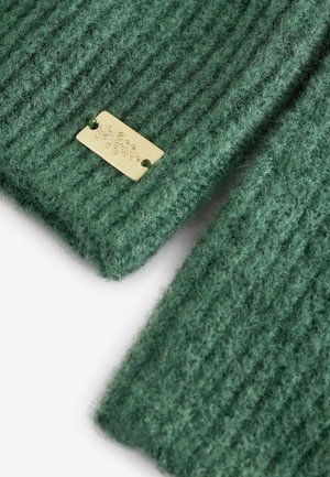 Green knitted fabric with ribbed texture, featuring a gold metal label attached, displaying engraved details.