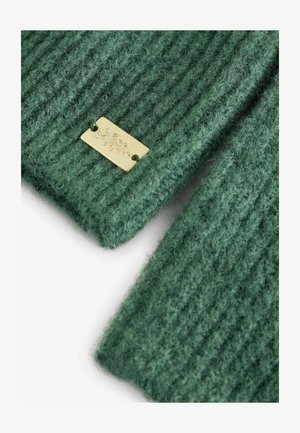 Green knitted fabric with ribbed texture, featuring a gold metal label attached, displaying engraved details.
