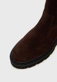 Botină din suede maro, cu vârful rotund, talpă ondulată neagră în contrast și fără hardware sau modele vizibile pe partea superioară.