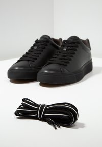 Baskets basses noires avec lacets noirs placées derrière une paire de lacets noirs enroulés avec des rayures blanches sur une surface blanche.