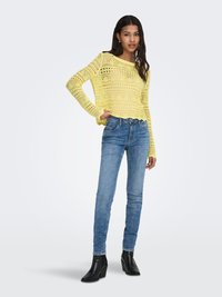 Vrouw draagt een gele, gehaakte top met lange mouwen, blauwe jeans en zwarte enkellaarzen met hak, staand met één hand op haar heup.