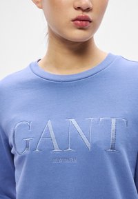 Felpa azzurro chiaro con logo "GANT" ricamato e scritta "NEW HAVEN". Tessuto morbido con scollo classico a girocollo. Design semplice e senza decorazioni.