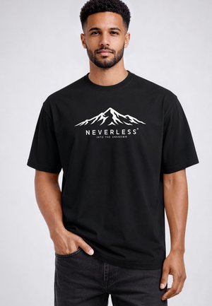 Mann trägt schwarzes T-Shirt mit weißem Bergmotiv und Text "NEVERLESS INTO THE UNKNOWN", steht mit einer Hand in der Tasche.