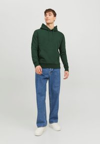 Felpa verde con tasca frontale, cappuccio con coulisse e polsini a coste. Abbinata a jeans blu larghi e sneakers bianche.