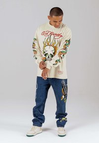 Langärmliges cremfarbenes Hemd mit Schädel- und Flammenmotiv, kombiniert mit blauen Jeanshosen, die Adler- und Schlangendesigns aufweisen. Weiße Sneaker.