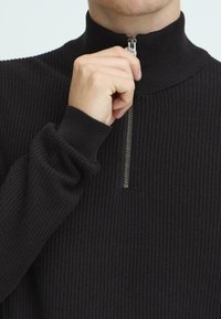 Maglione lavorato a maglia nero con colletto alto e design a mezza zip. Caratterizzato da una texture a coste e polsini aderenti. Nessun motivo o dettaglio aggiuntivo.
