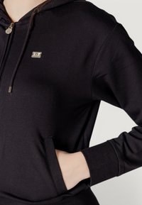 Felpa nera con zip realizzata in tessuto morbido, dotata di una tasca frontale, polsini a costine e un accento del logo metallico sul petto.