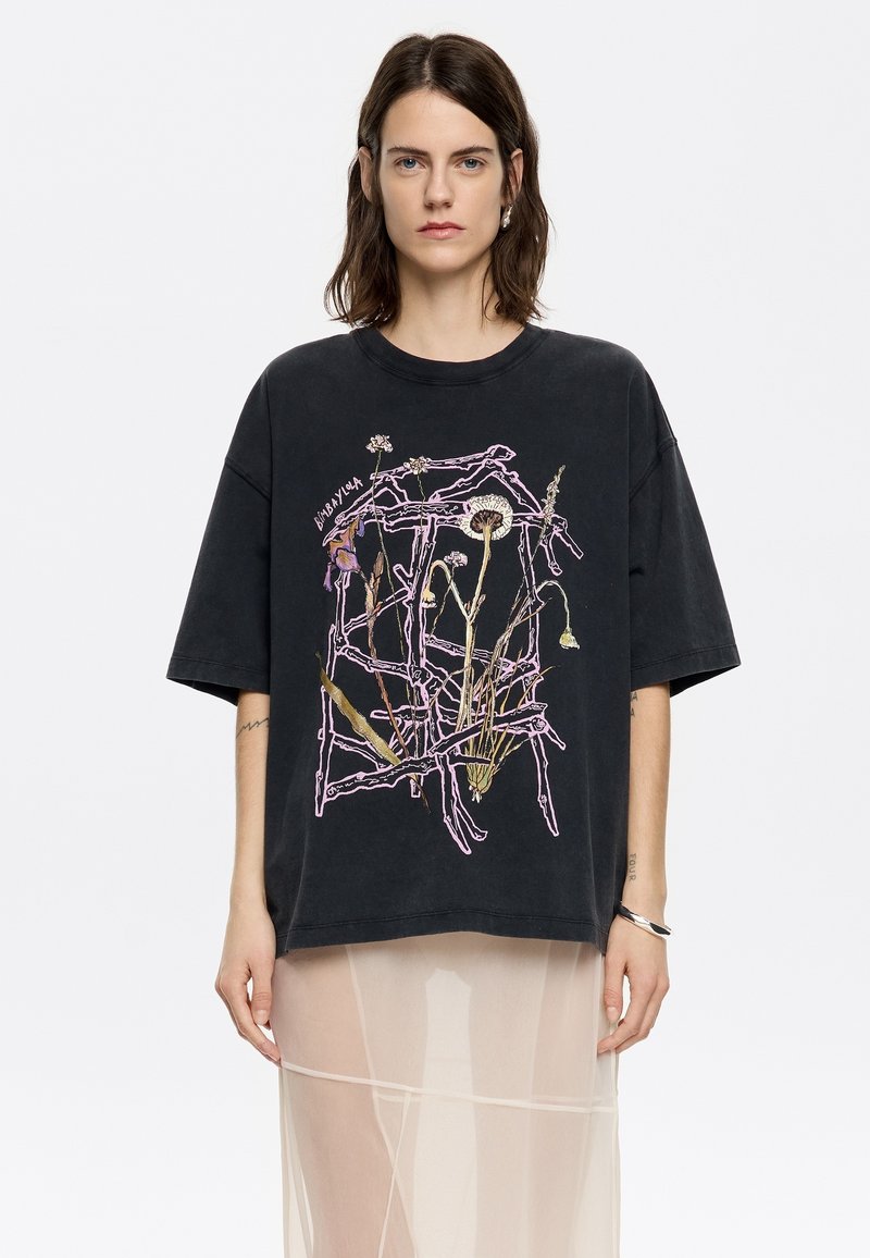 Juodas per didelis t-shirt su gėlių grafika purpurine ir kremine spalvomis, su apvaliu kaklu ir trumpomis rankovėmis. Dizaino elemente įtraukta tekstas.
