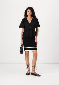 Svart peplum-overdel med blonder og knapper, kombinert med et svart ribbestrikket blyantskjørt med en hvit stripe. Sorte flate sko og håndveske.
